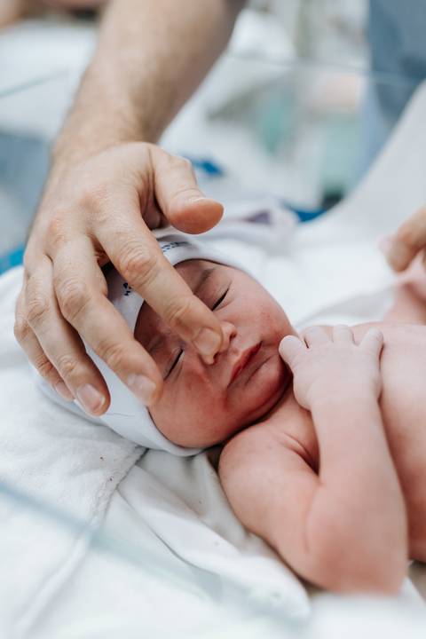 Foto em detalhe do pai fazendo carinho no rosto do recém-nascido.'