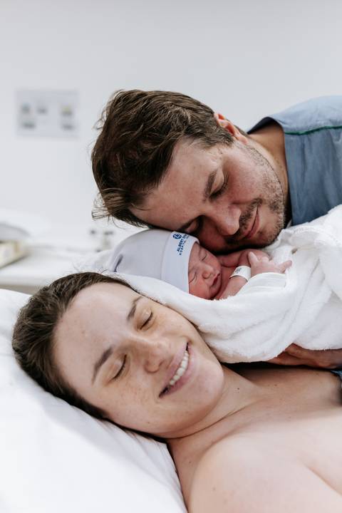 Retrato do casal sorrindo com o bebê adormecido entre eles.'