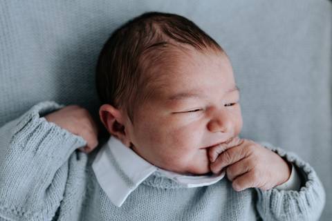 Close do Guilherme com a mãozinha no rosto usando conjunto de tricô azul.'