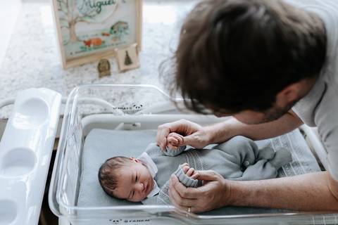 Pai interagindo com o filho no berço de acrílico da maternidade.'