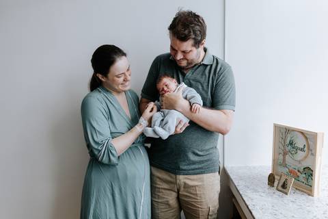 Retrato da família no quarto do hospital: pai segurando o bebê ao lado da mãe.'