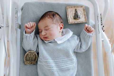 Foto do bebê Miguel dormindo no berço da maternidade, usando conjunto de tricô azul e rodeado por medalhas religiosas de proteção.'