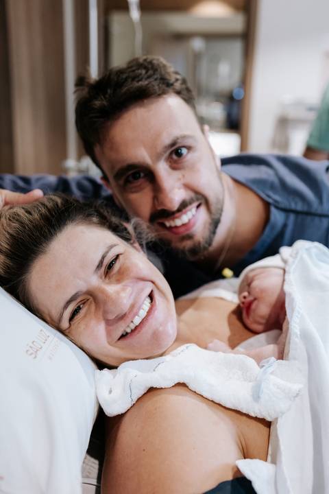 Foto do casal radiante com a bebê no pós-parto imediato.'