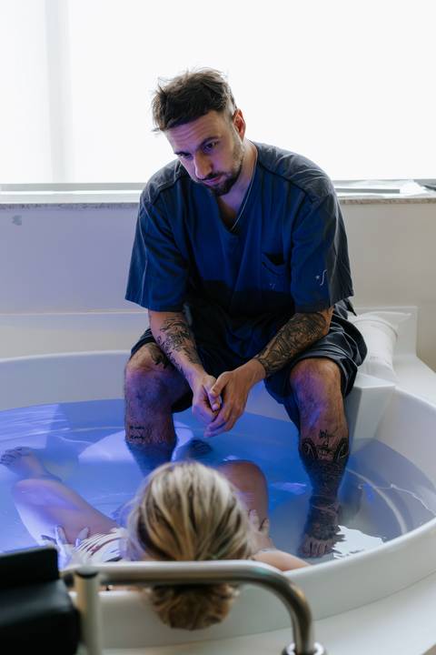 Foto do parceiro acompanhando o parto à beira da banheira.'