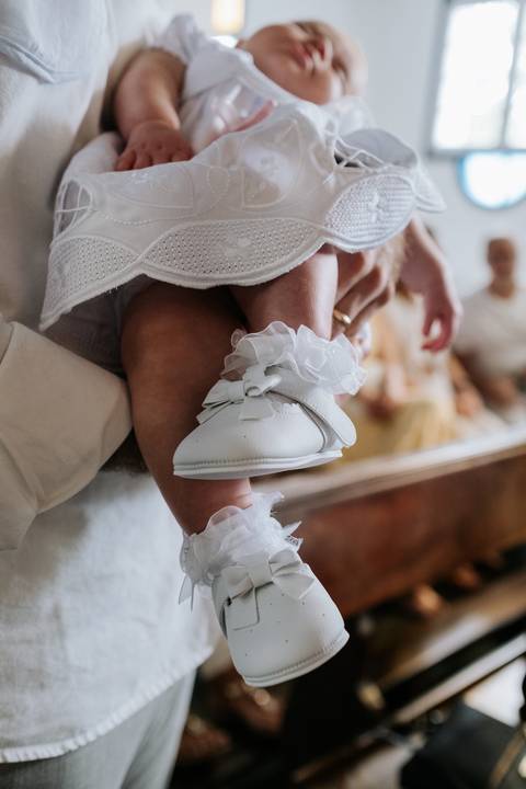 Foto em detalhe do sapatinho branco de laço e das meias com babados da bebê.'