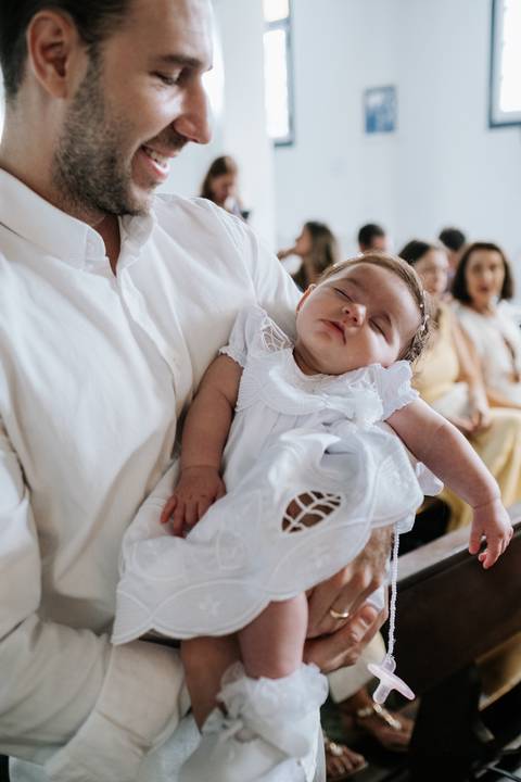 Foto da bebê Olívia dormindo profundamente no colo durante a cerimônia.'