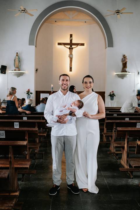 Foto do casal sorrindo com a bebê nos bancos da igreja.'