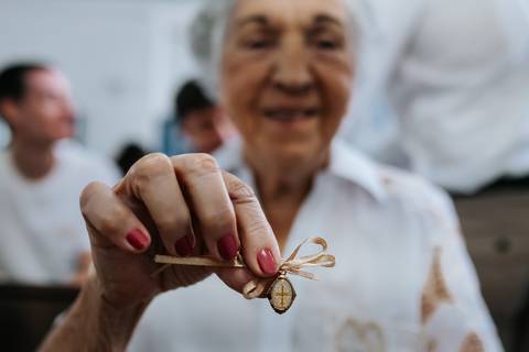 Foto em detalhe de uma medalhinha religiosa de batismo sendo segurada pela bisavó. '