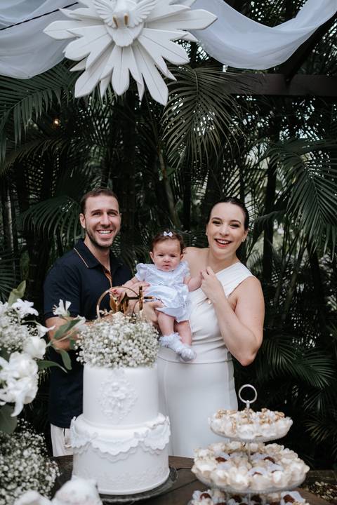 Foto dos pais sorridentes com a bebê Olívia na mesa do batizado.'