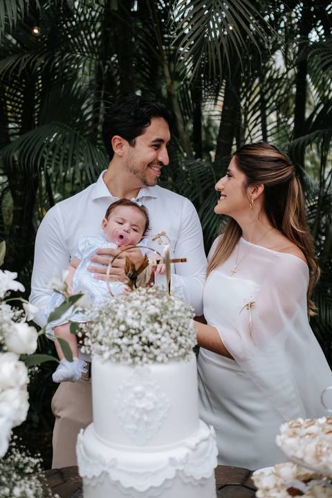 Foto dos padrinhos sorrindo com a bebê durante a recepção.'