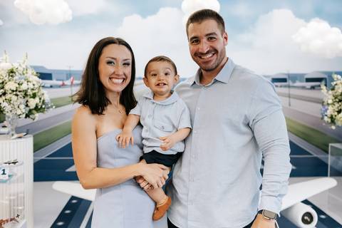Foto dos pais sorridentes com o aniversariante no colo em frente a mesa principal.'