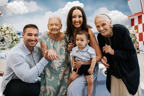 Foto da família completa, incluindo avós, posando na mesa do bolo.'
