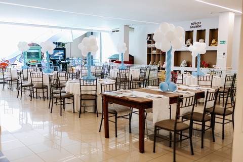 Foto panorâmica da sala de festa com mesas e cadeiras decoradas.'