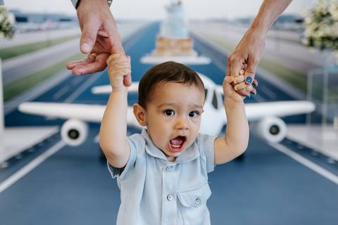 Foto do aniversariante sorrindo enquanto é guiado por mãos adultas.'