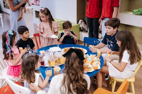 Foto aproximada de crianças reunidas lanchando na mesa da festa.'
