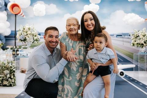 Foto dos pais e avó sorridentes com o aniversariante na mesa do bolo.'