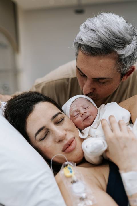 Foto do pai beijando a cabeça do bebê junto com a mãe.'