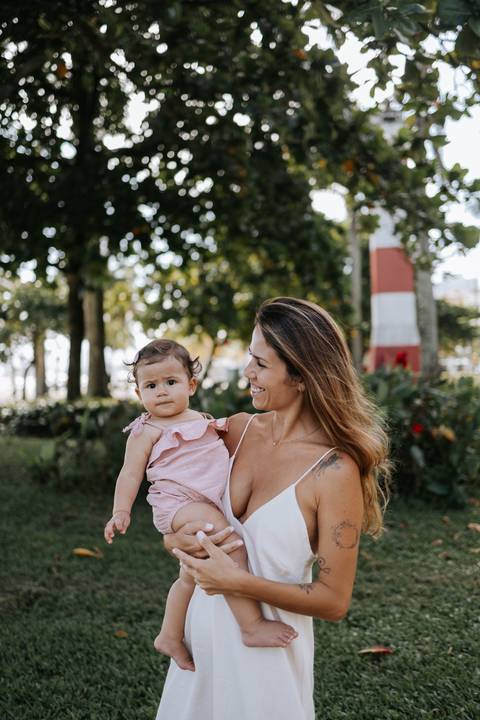 Foto a mãe segurando a filha no colo em um gramado com coqueiros.'