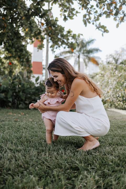 Foto da mãe agachada no gramado brincando com a filha.'