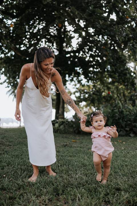 Foto da mãe caminhando de mãos dadas com a filha que está aprendendo a andar.'