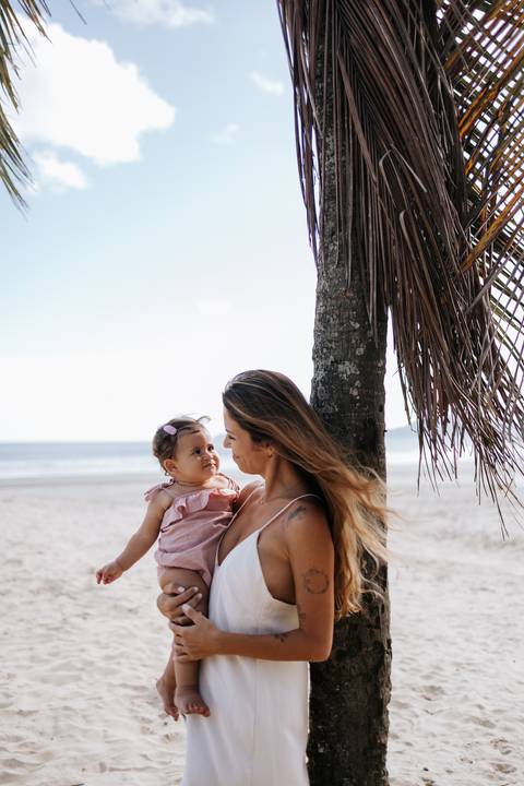 Foto da mãe e filha sentadas na areia da praia encostadas em um coqueiro.'