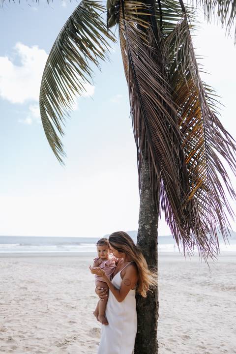 Foto panorâmica da praia com mãe e filha em destaque sob um coqueiro.'