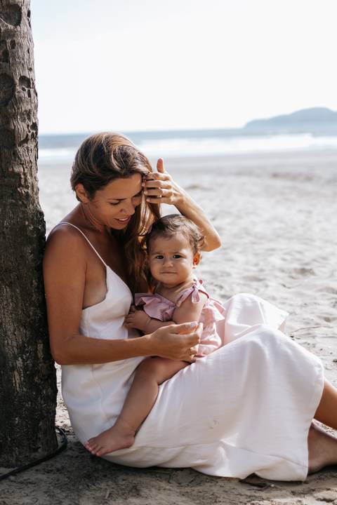 Foto da mãe sentada na areia abraçando a filha com o mar ao fundo.'