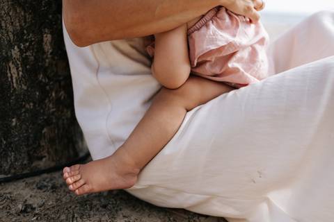 Foto em detalhe das pernas e pés da bebê no colo da mãe na areia.'