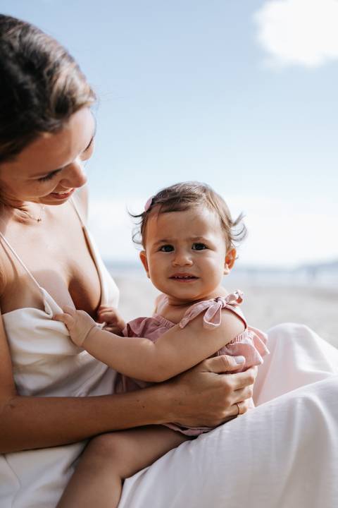 Foto aproximada da bebê no colo da mãe com o mar ao fundo.'