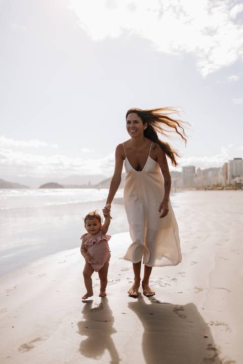 Foto da mãe caminhando de mãos dadas com a filha na beira da água.'