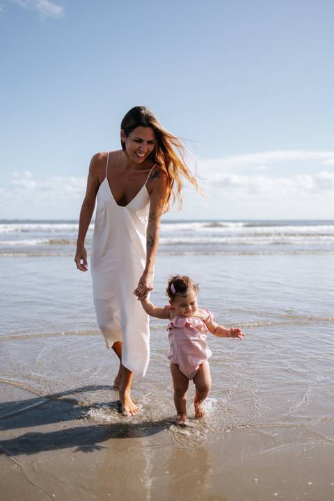 Foto da mãe sorridente guiando os primeiros passos da filha na praia.'