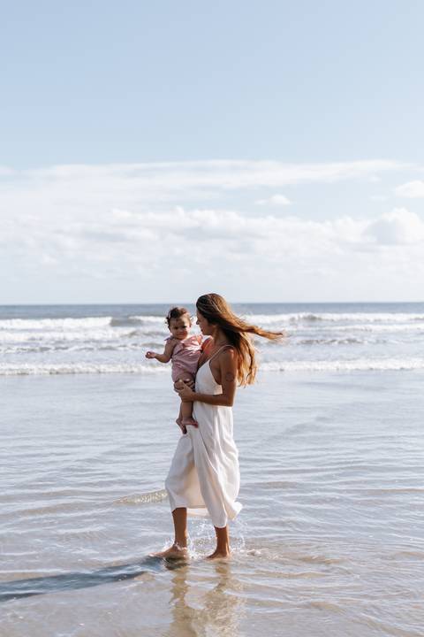Foto da mãe de perfil segurando a filha no colo diante das ondas.'