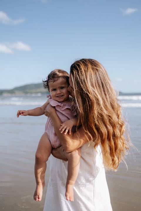 Foto da mãe beijando o rosto da pequena à beira-mar.'