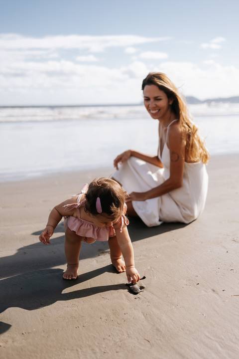 Foto da bebê engatinhando na areia enquanto a mãe observa ao fundo.'