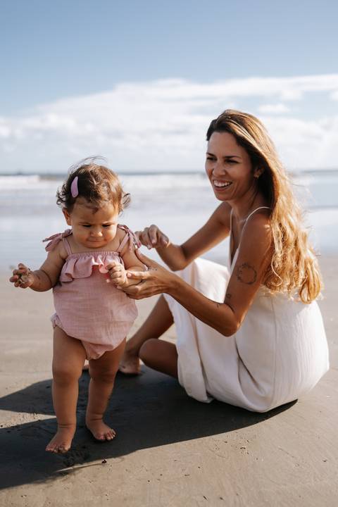 Foto da mãe sentada na areia brincando com as mãos da filha.'