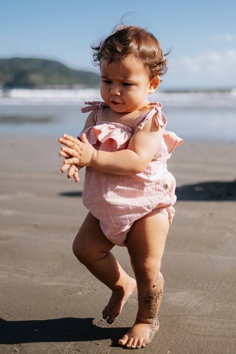 Foto aproximada da bebê em pé na areia com seu romper rosa.'