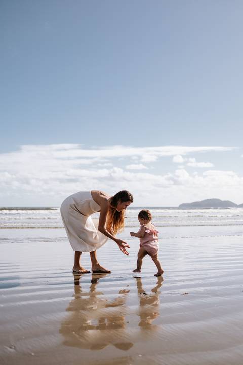 Foto da mãe agachada na beira-mar chamando a bebê para caminhar.'