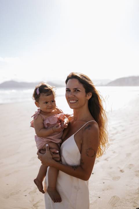 Foto da mãe sorridente segurando a filha no colo diante do mar.'