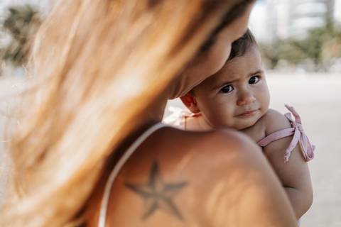 Foto de close da bebê apoiada no ombro da mãe, olhando para a câmera.'