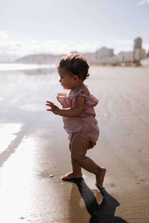 Foto de perfil da bebê em pé na beira da água sob a luz do sol.'