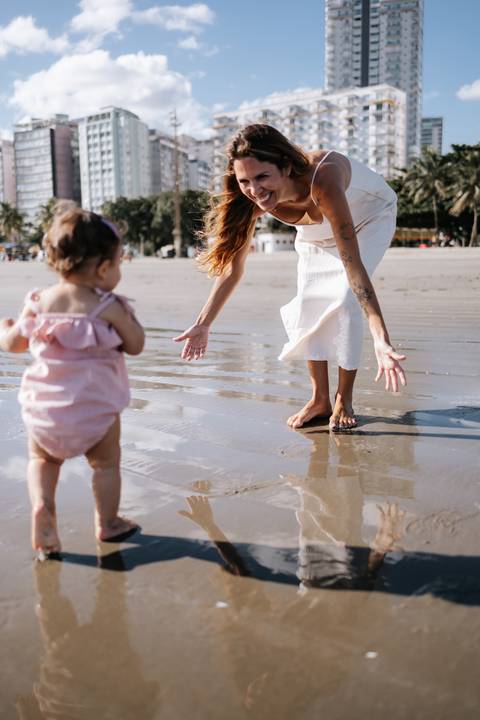 Foto da mãe e filha brincando com o reflexo da água na areia molhada.'