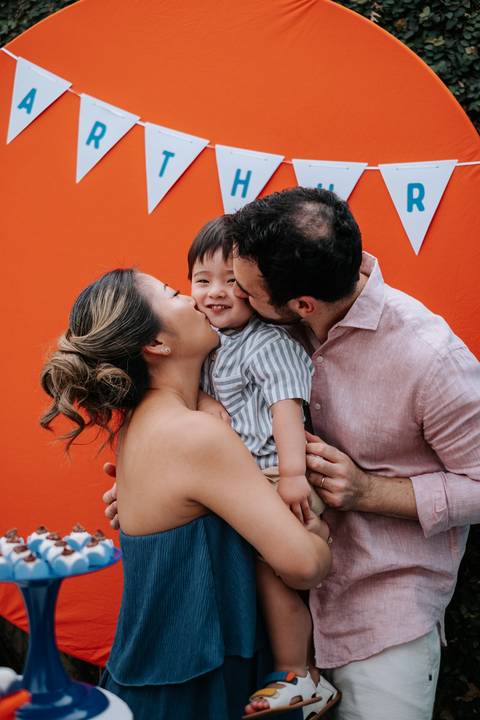 Foto da mãe e do pai beijando o aniversariante ao mesmo tempo.'