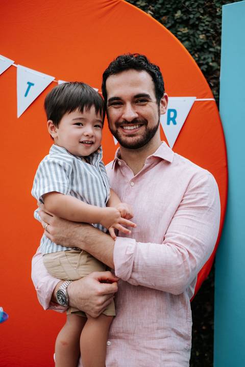 Foto do pai sorridente segurando o Arthur em frente ao painel laranja.'