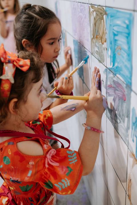 Foto em detalhe de convidada mirim pintando com pincel no painel de azulejos.'