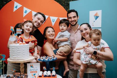 Foto da família completa e amigos celebrando ao redor do bolo.'
