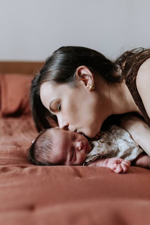 Foto da mãe beijando delicadamente a testa da bebê na cama.'