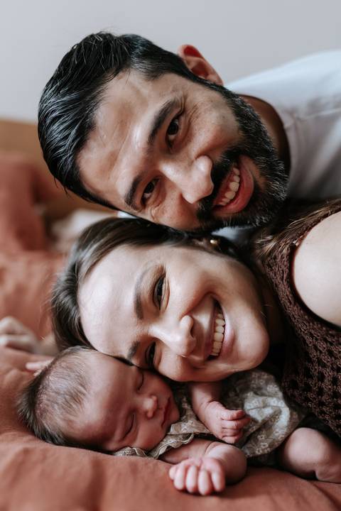 Foto do casal sorridente deitados com a Olívia entre eles.'