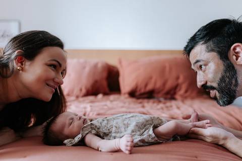 Foto do casal trocando olhares sobre a Olívia que descansa na cama.'