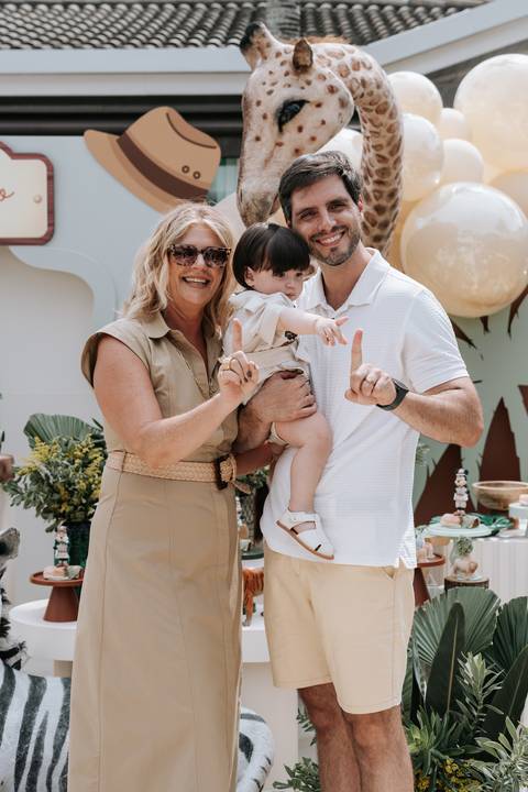Foto do aniversariante com os avós fazendo o número 1 com as mãos.'