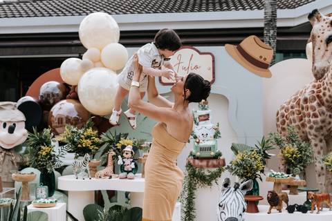 Foto da mãe erguendo o aniversariante para o alto em frente à mesa principal.'
